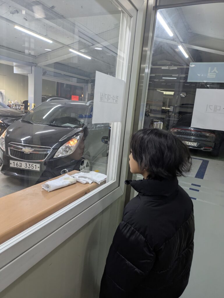 어두운 정비소에서 유리창을 통해 바깥을 바라보는 아이의 모습(차량 정기 검사)